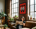 Person reading a book on a brown couch in a room with large windows and a red poster on the wall.